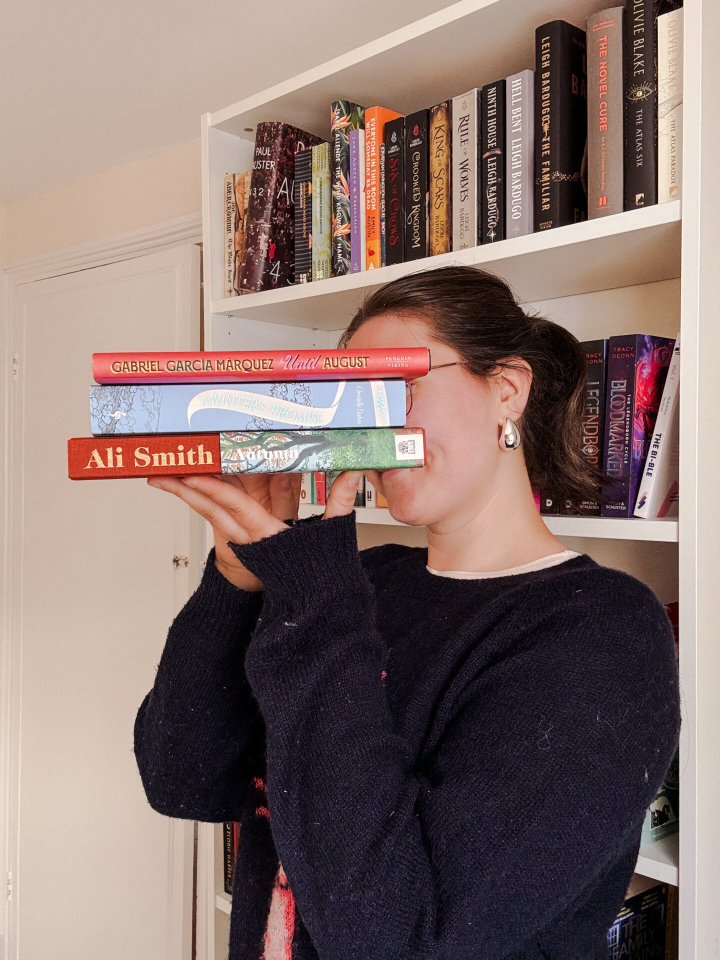 Girl in a blue jumper standing in front of bookshelves and holding a stack of book sin front of her face